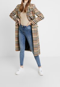 Houndstooth coat in beige, brown, and multicolor; fitted light sweater; blue skinny jeans; white sneakers; smooth texture and tailored design.