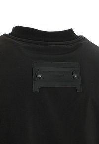 Chaqueta negra por detrás con cuello de canalé y un parche rectangular cosido asegurado por dos botones cerca del escote.