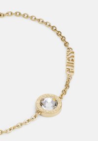 Bracelet en chaîne dorée avec un pendentif circulaire orné d'une pierre claire et facettée. Le pendentif est gravé de "GUESS" sur son bord.