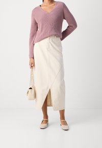Pullover rosa chiaro a maglia con scollo a V, abbinato a una gonna avvolgente beige a vita alta e a delle scarpe beige slip-on. Piccola borsa a mano beige.