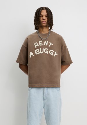 Hnedé oversized tričko s potlačou "RENT A BUGGY" v off-white farbe. Krátke rukávy, okrúhly výstrih, mäkký bavlnený materiál, voľný strih.