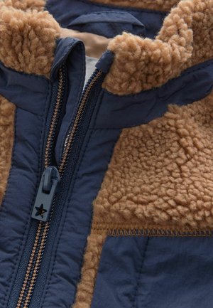Jachetă albastru marin cu un guler pufos din blană maro, care are un fermoar cu o agățătoare în formă de stea și detalii de cusătură contrastante.