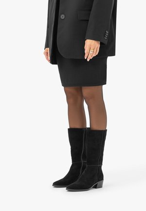 Femme portant des bottes en daim noires montant jusqu'aux genoux, des collants noirs transparents, une jupe noire et un blazer noir avec des bagues aux doigts.
