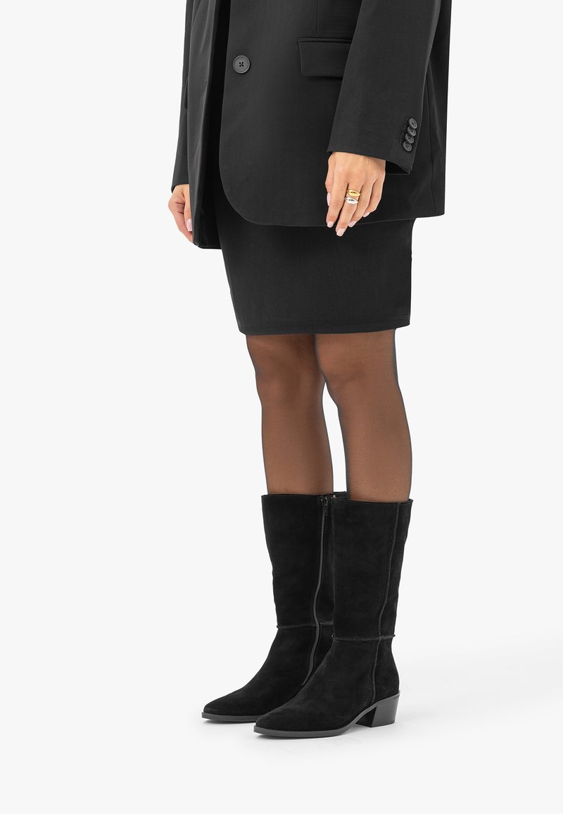 Femme portant des bottes en daim noires montant jusqu'aux genoux, des collants noirs transparents, une jupe noire et un blazer noir avec des bagues aux doigts.
