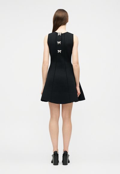 Γυναίκα που φοράει μαύρο φόρεμα χωρίς μανίκια με τρία μικρά λευκά φιογκάκια στην πλάτη, συνδυασμένο με μαύρα παπούτσια με χοντρό τακούνι.