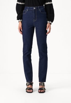 Vrouw dragen donkerblauwe slim-fit jeans, zwarte blouse met lange mouwen met witte kanten details en zwarte sandalen met gouden gespen.