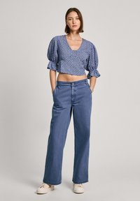 Blauwe gebloemde crop top met pofmouwen, gecombineerd met hoge taille blauwe jeans. Model staat met handen in de zakken en draagt lichtere schoenen.
