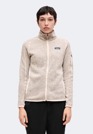 Persoon met kort donker haar, een beige fleecejack met rits en een zwarte broek, staand tegen een effen lichte achtergrond.
