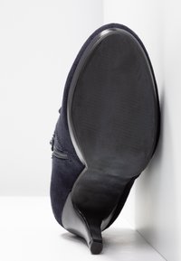 Zapato de tacón alto de ante azul marino, colocado verticalmente sobre un fondo claro, mostrando los detalles de la suela y el tacón.