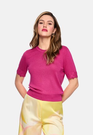 Frau mit langen braunen Haaren, die ein fuchsiafarbenes kurzärmeliges Oberteil mit gezackten Kanten und hellgelbe Hosen mit abstrakten Mustern trägt.