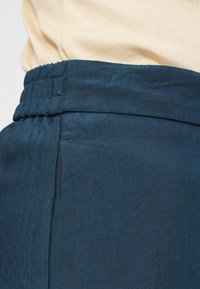 Kalhoty z hladkého materiálu v námořnické modři s elastickým pasem a bočními švy. Mají jemný lesk a jsou střihově přizpůsobené.