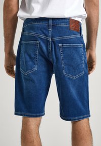 Pantalones cortos de mezclilla azul con un ajuste estándar, que presentan una etiqueta de marca de cuero, dos bolsillos traseros y costuras de contraste a lo largo de los bordes.