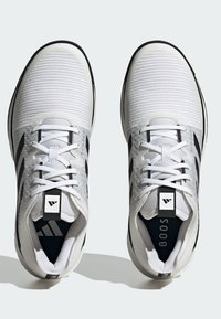 Weiße Sportschuhe mit gerippter Struktur, schwarzen Akzenten, Schnürdesign, Logo auf der Zunge und gepolstertem Innenfutter für Komfort.
