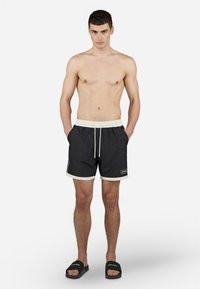 Shorts de bain noirs avec cordon blanc et poignets, présentant des rayures latérales et un patch de logo. Tissu lisse avec une coupe décontractée.
