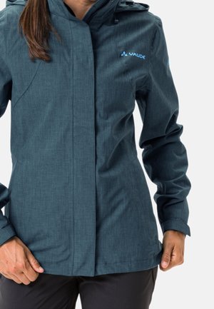 Kobieta nosząca ciemnogranatową wodoodporną kurtkę z kapturem, przedni zamek przykryty zakładką przeciwdeszczową oraz logo Vaude na piersi.