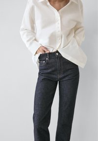 Camicia bianca con bottoni, vestibilità comoda, maniche lunghe. Jeans in denim scuro, gamba dritta, design classico con cinque tasche. La texture appare morbida e nitida.