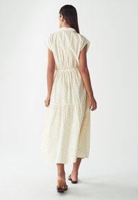 Šaty krémové barvy s krátkými rukávy a černými puntíky, se zúženým pasem a střihem do více vrstev, sahajícími do poloviny lýtek. Hladká látka s límečkem.