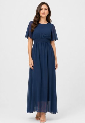 Vrouw draagt een marineblauwe maxi-jurk met wapperende mouwen en een gerimpelde taille, staand tegen een effen lichte achtergrond.