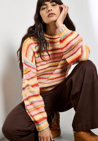 Sweter z wielokolorowym paskiem, w odcieniach pomarańczowym, różowym i niebieskim. Luźny krój, ściągacze przy mankietach i okrągły dekolt, w połączeniu z brązowymi spodniami.
