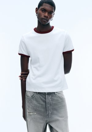 Jeune homme portant un t-shirt blanc avec col et manches rouges, et un jean gris déchiré, debout devant un fond clair uni.