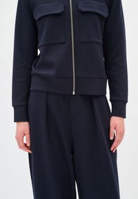 Giacca con zip blu navy con due tasche sul petto, realizzata in tessuto liscio. Abbinata a pantaloni blu navy a vestibilità ampia con una leggera texture.