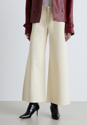 Person trägt cremefarbene Hosen mit weitem Bein, schwarze spitze Stiefel mit Absatz, eine bordeauxrote strukturierte Jacke und Ringe an beiden Händen vor einem einfarbigen Hintergrund.