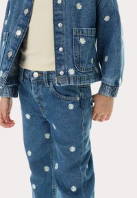 Conjunto de chaqueta y jeans de mezclilla, azul con bordado de margaritas blancas, que presenta botones plateados, textura suave y puños ajustados.
