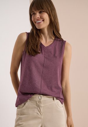 Femme souriante portant un haut sans manches violet à rayures avec un col en V et un pantalon beige clair sur un fond neutre.