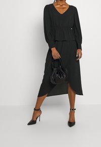 Schwarzes langärmliges Kleid mit V-Ausschnitt und Peplum-Taille, versehen mit einem High-Low-Saum. Kombiniert mit schwarzen Stilettos und einer kleinen schwarzen Tasche.