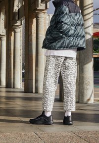 Giubbotto imbottito in verde scuro con un motivo a trama, abbinato a pantaloni bianchi con una stampa grafica nera e scarpe da ginnastica nere.