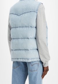 Gilet imbottito in denim azzurro con collo alto e dettagli cuciti, indossato sopra una maglia grigia a maniche lunghe, abbinato a jeans blu.