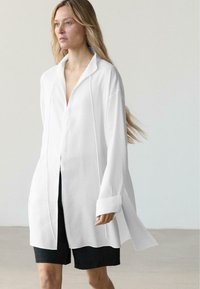 Kvinde med langt blondt hår iført en stor hvid skjorte med knapper og sorte knælange shorts foran en ensartet lyse baggrund.