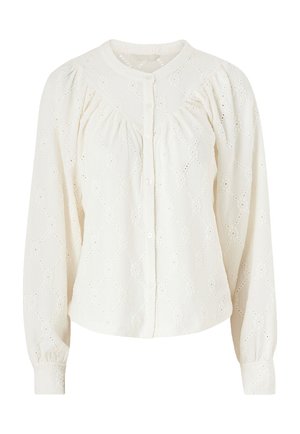 Blouse couleur crème avec un motif en losanges, manches longues bouffantes, col rond et fermeture à boutons. Tissu texturé avec des détails en œillets.