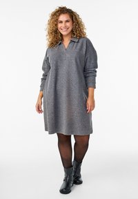 Kobieta z kręconymi włosami, ubrana w szaro-długą sukienkę z długim rękawem, czarne rajstopy i czarne botki, uśmiechająca się i idąca do przodu na białym tle.