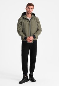 Giacca a hood verde oliva con zip frontale, polsini e orlo neri, abbinata a pantaloni jogger neri e sneakers alte nere, in piedi su uno sfondo bianco.