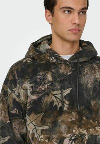 Hoodie en polaire arborant un motif de camouflage sombre avec des feuilles dans des teintes de marron et de vert, doté d'une capuche avec cordon de serrage.