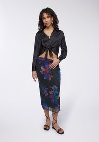 Camicia nera in raso corta con bottoni e dettaglio a cravatta, abbinata a una gonna midi trasparente nera con stampa floreale e sandali neri con cinturino.