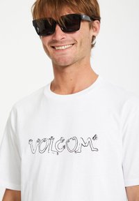 T-shirt en coton blanc avec un logo graphique noir et des motifs de mains abstraits. Encolure ronde, manches courtes et coupe décontractée.