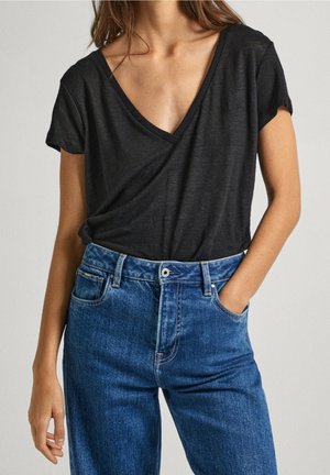 Kobieta ubrana w czarną bluzkę z krótkim rękawem i dekoltem w serek, wpuszczoną do wysokich niebieskich dżinsów, z jedną ręką w kieszeni.