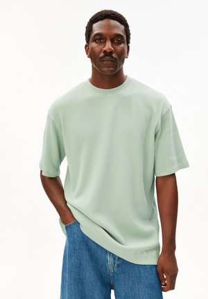 Mann mit kurzen lockigen Haaren und Schnurrbart, trägt ein hellgrünes, strukturiertes Kurzarmhemd und blaue Jeans, steht mit einer Hand in der Tasche.