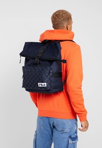 Námornícky modrý batoh FILA s dynamickým překlopným víkem, texturovanou spodní kapsou a nastavitelným popruhem. Obsahuje logo a klipsov é uzavření.