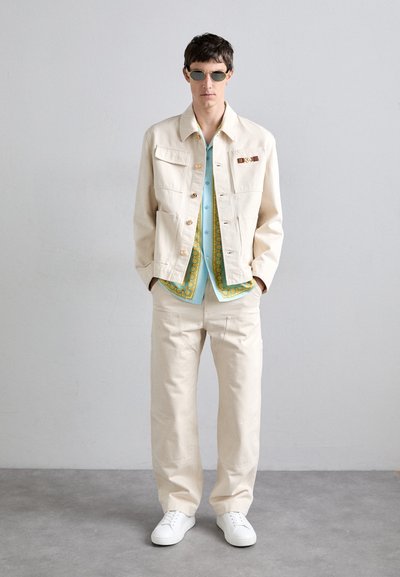 Beige denim jacket with gold hardware, paired with a light blue shirt featuring a green paisley pattern, and cream trousers with white sneakers.