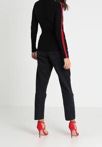 Svart ribbad tröja med röd rand, matchad med svarta högmidjade jeans och röda högklackade sandaler. Bakifrån visad hållning.