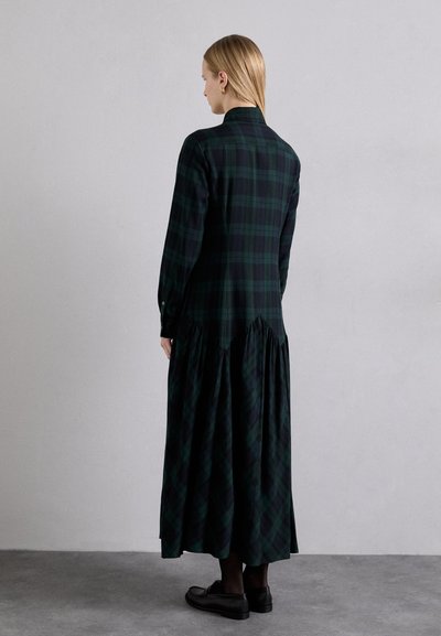 Long-sleeve plaid dress in dark green and black, featuring a fitted bodice, gathered waist, and flowing skirt. Black shoes complement the outfit.