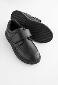 Black leather children's shoes with wide velcro straps and rubber soles, one shoe upright and the other showing the tread pattern.