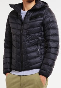 Svart pufferjacka med huva, med kviltat mönster, dragkedjefickor och en slät, glänsande textur. Stängning med dragkedja fram.