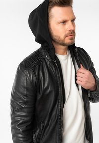 Giubbotto da motociclista in pelle nera con dettagli a coste sulle spalle, chiusura con zip e un cappuccio nero indossato sotto. Texture liscia, design aderente.