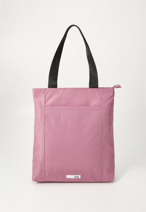 Sac fourre-tout rose clair en nylon lisse, avec des poignées noires portant un logo et une poche avant. Détail zippé en haut.