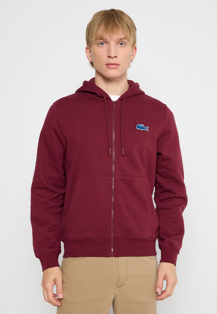 Sweat à zip marron en tissu côtelé, avec une poche kangourou et un petit logo bleu. Contient une capuche avec cordon de serrage. Texture lisse.