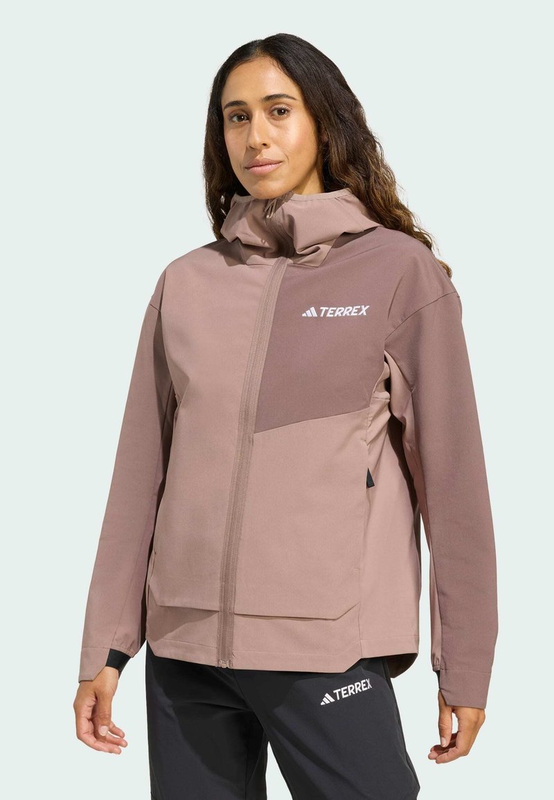 Veste légère en shell doux dans des teintes de rose tendre, avec un col haut, une fermeture éclair à l'avant et un détail de logo. Le matériau a une texture lisse.